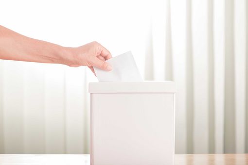 In the centre of the picture is a white ballot box in the shape of a square box. A hand is placing its ballot paper in the ballot box.
