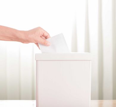In der Mitte des Bildes steht eine weiße Wahlurne in Form einer quadratischen Box. In die Wahlurne legt eine Hand ihren Stimmzettel ab.