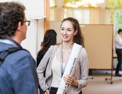 Zwei Menchen im Gespräch in der Villa Tillanns, eine Person von hinten zu sehen weiter vorne im Bild, eine Person mit Poster in der Hand von vorne zu sehen