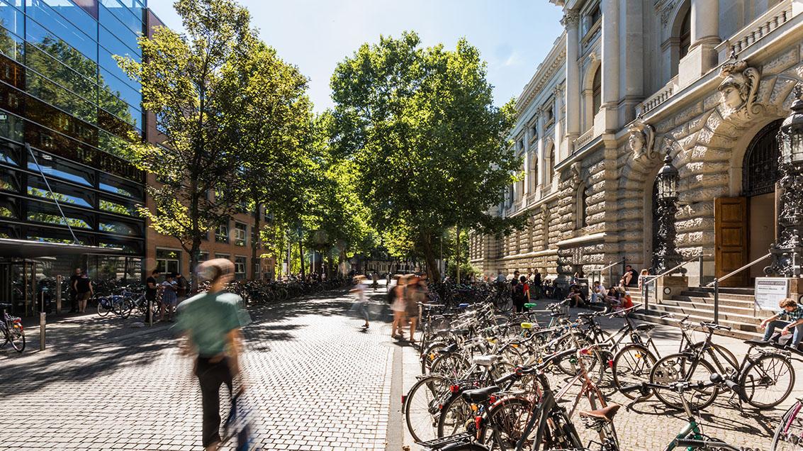 zur Vergrößerungsansicht des Bildes: Studierende vor der Universitätsbibliothek. Ihre Umrisse sind unscharf, da sie in Bewegung sind