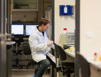 Foto: Ein Student der Biowissenschaften in einem Kittel gekleidet und mit blauen Füßlingen sitzt am Schreibtisch eines Labors
